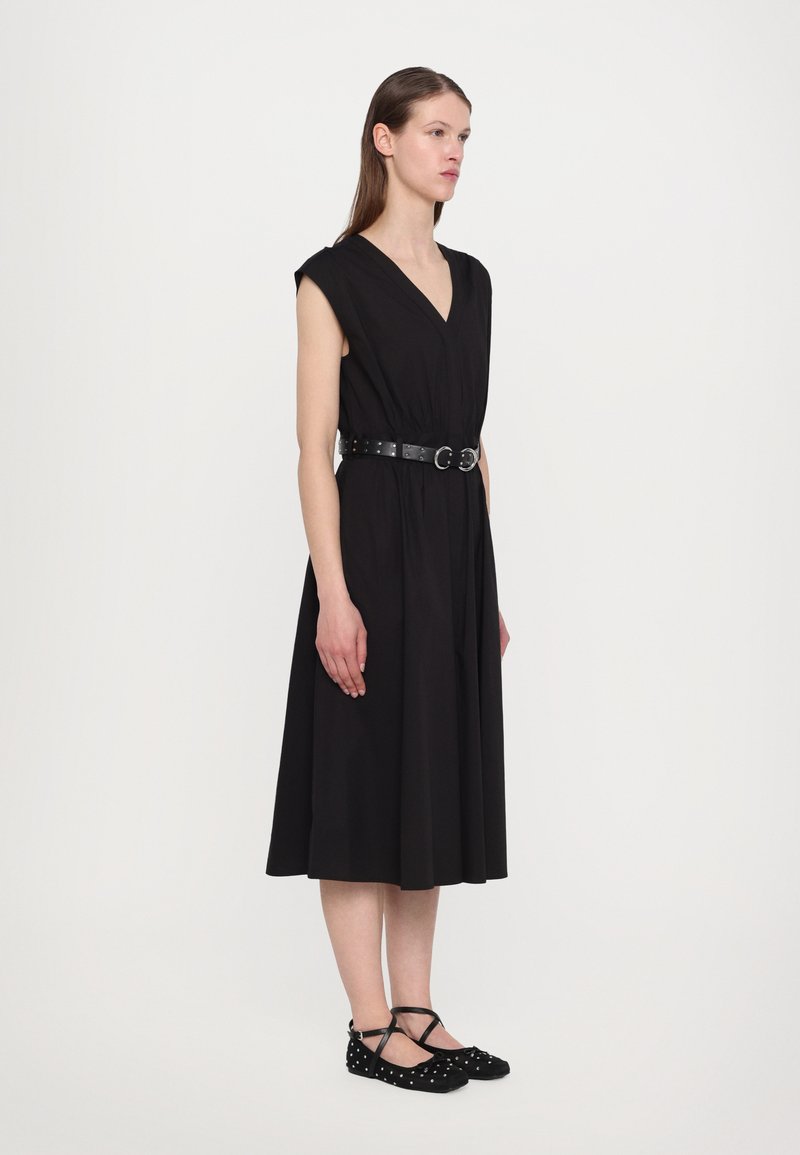 Vestido negro sin mangas con un escote en V profundo y una cinturilla, que cuenta con una falda acampanada. Se lleva con unos zapatos planos negros adornados con lunares blancos.