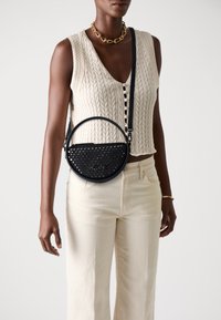 Black, round crossbody bag with perforated design and metallic accents, featuring a zipper closure and adjustable strap. Worn with a cream knit top.