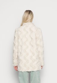 Chaqueta larga de piel sintética color crema con textura ondulada. Presenta un cuello de chal y hombros caídos, ideal para superponer sobre varios conjuntos.