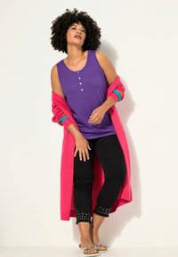 Canottiera viola con bottoni e tasca, abbinata a un lungo cardigan rosa brillante. Pantaloni neri a cropped con orlo borchiato e sandali argentati.