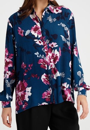 Femme portant un chemisier bleu à manches longues boutonné avec un motif floral rose, blanc et noir, associé à un pantalon noir.
