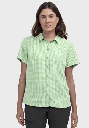 Frau trägt ein kurzärmeliges hellgrünes Vichy-Karoshirt und schwarze Hose und steht vor einem schlichten weißen Hintergrund.