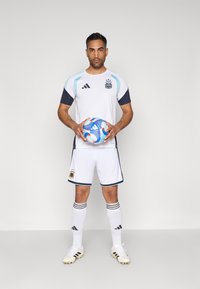 Λευκή ποδοσφαιρική φανέλα με ανοιχτό μπλε λεπτομέρειες, ναυτικές μανσέτες, αντίστοιχες βερμούδες και μαύρες ρίγες στις κάλτσες, κρατώντας μια ποδοσφαιρική μπάλα με μπλε και κόκκινο σχέδιο.