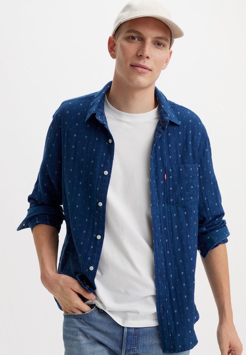 Camicia blu con bottoni e taschino, caratterizzata da una trama a motivo. Indossata sopra una maglietta bianca, abbinata a jeans azzurri chiari e un cappellino.