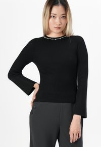 Young woman with straight blonde hair wearing a black ribbed long-sleeve top with pearl detail at neckline and dark high-waist pants.