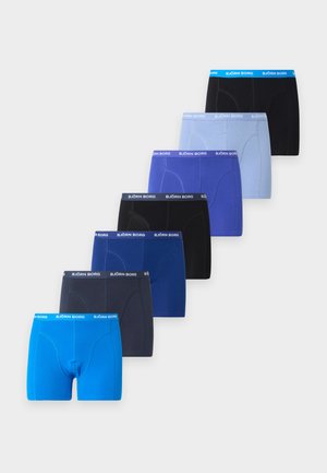 Uomo magro dalla pelle scura in piedi a piedi nudi, indossa boxer azzurri chiari con fascia in vita marchiata, guarda leggermente verso destra su uno sfondo semplice.