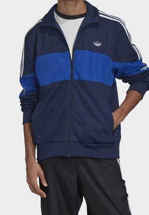 Mann trägt eine marineblaue und blaue Trainingsjacke mit weißen Streifen an den Ärmeln, kombiniert mit schwarzen Hosen, Hand in der Tasche.