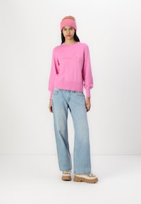 Pull rose avec "voilà" brodé en écriture cursive, associé à un jean ample bleu clair et des chaussures à semelle épaisse beige. Bonnet tricoté multicolore.