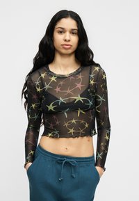 Νεαρή γυναίκα με μακριά σκουρόχρωμα μαλλιά, που φοράει μαύρο διαφανές crop top με μακριά μανίκια και πολύχρωμο μοτίβο αστεριών, καθώς και μπλε παντελόνι με κορδόνι στη μέση.