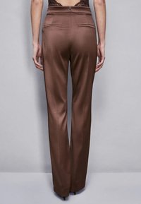 Pantaloni marroni, su misura, con una texture liscia, gamba dritta e due tasche posteriori; chiusura con zip in vita.
