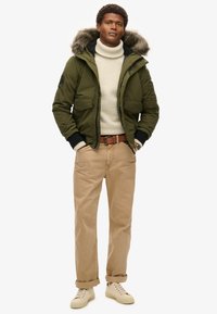Parka verde con bordo in pelliccia, chiusura con zip e bottoni a pressione, due tasche frontali, abbinata a un dolcevita crema, pantaloni beige e sneakers chiare.