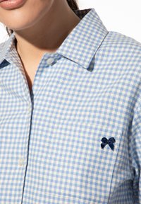 Hellblau und weiß kariertes Hemd mit Kragen, versehen mit einer blauen gestickten Schleife auf der Brust, aus Baumwollstoff gefertigt.