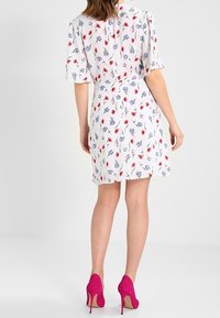 Kvinna iförd en vit blommig klänning med röda och blå blommor, kombinerad med klara rosa högklackade skor, stående med ryggen vänd.