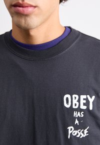 Tricou din bumbac negru cu text grafic alb "OBEY HAS A POSSE" pe față. Guler rotund cu o căptușeală interioară mov. Textură moale.
