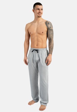 Oberkörperfreier Mann mit detailliertem Vollarm-Tattoo am rechten Arm, trägt grau-weiß gestreifte Hosen mit vertikalen Streifen, steht barfuß, schaut nach rechts und lächelt.