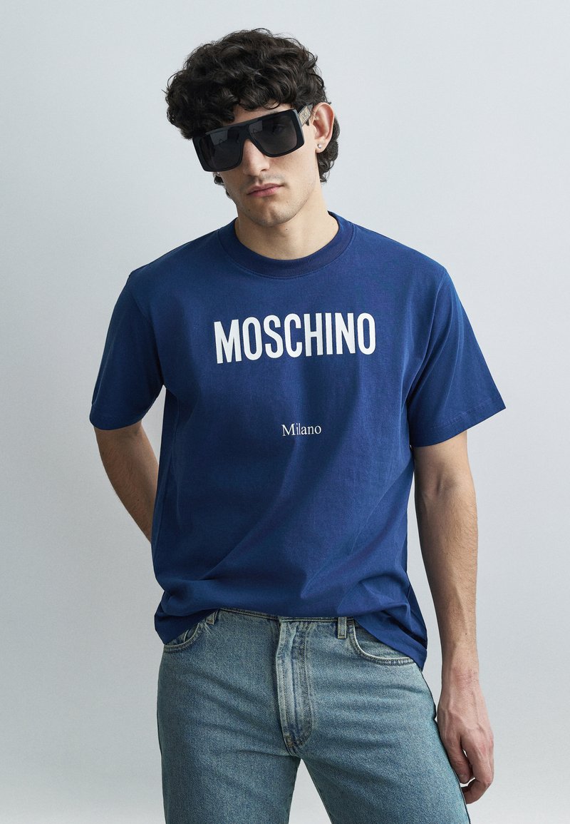 Blauw katoenen T-shirt met "MOSCHINO" en "Milano" in witte letters, gecombineerd met lichtblauwe spijkerbroek en oversized zwarte zonnebril.