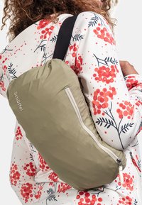 Khakifarbener Rucksack mit runder Form, ausgestattet mit einem Reißverschluss und einem Logo. Wird über einem weißen, gemusterten Hemd mit roten Blumendetails getragen.