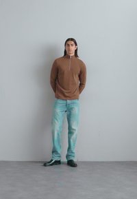 Brauner Zip-Pullover, hellblaue ausgewaschene Jeans, schwarze Schuhe. Modell steht mit den Händen hinter dem Rücken an einer grauen Wand.