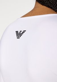 Camiseta atlética branca com textura lisa, apresentando um logótipo preto com riscas horizontais na parte superior das costas.