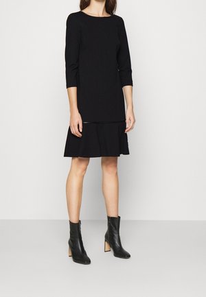 Mujer con un vestido negro hasta la rodilla con mangas tres cuartos y botines negros con tacón, posando frente a un fondo claro y liso.