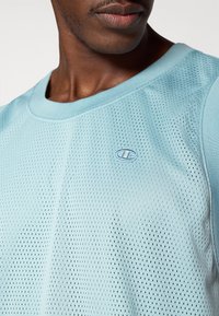 Αθλητική μπλούζα ανοιχτού γαλάζιου χρώματος με λαιμόκοψη, με μικρό κεντητό λογότυπο. Η υφή είναι αν απνεύσιμη με πουά σχέδιο.