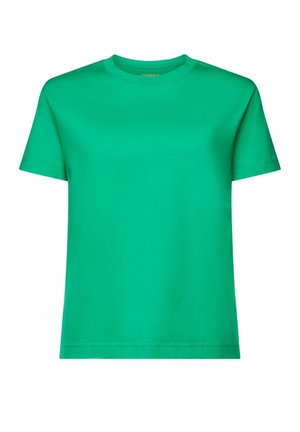 Person iført en knaldgrøn skjorte, sorte bukser, en sort-hvid stribet sweater bundet omkring skuldrene og bærende en lille grøn håndtaske.