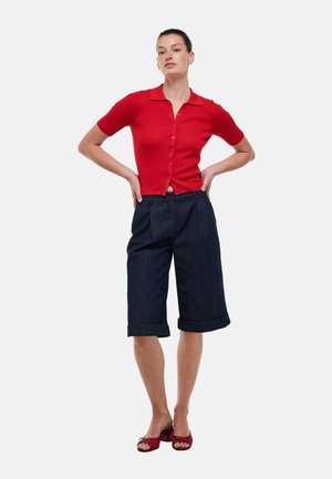 Femme debout, les mains sur les hanches, portant un haut rouge à manches courtes boutonné, un short en jean foncé au-dessus du genou et des escarpins rouges à bout ouvert.