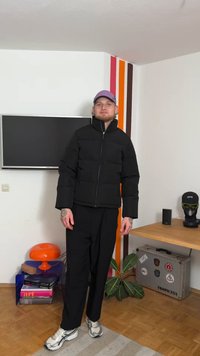 Jaqueta preta acolchoada com gola alta e fecho de correr frontal. Combinada com calças pretas e sapatilhas prateadas. O quarto possui uma televisão e plantas decorativas.