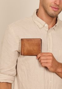 Brown leather wallet held by a person, featuring a smooth texture and a simple design. The wallet is folded with a subtle embossed logo.