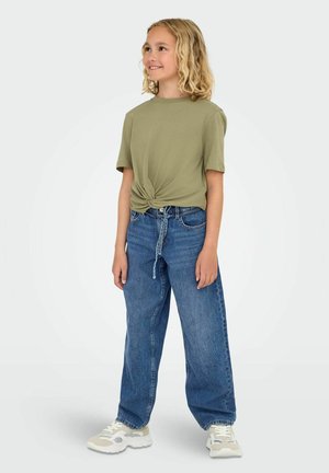 T-shirt olive vert avec nœud devant, jeans en denim bleu amples, et baskets blanches avec accents gris, se tenant devant un fond clair.