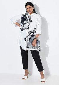 Long white blouse with black floral print, three-quarter sleeves, paired with black fitted pants and white open-toe heels. Embellished clutch bag.