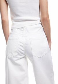 Valkoiset denimit farkut, joissa on rento leikkaus. Niissä on kaksi takataskua, joissa on kulmikkaat läpät ja ommeltu yksityiskohtia. Pintamateriaali on sileä ja kevyen näköinen.