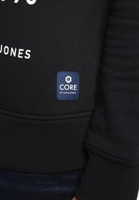 Sudadera negra con texto blanco, puños y dobladillo acanalados, y una etiqueta azul etiquetada "CORE by JACK & JONES" en la parte inferior izquierda delantera.