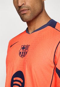 Élénk narancssárga labdarúgó mez tengerészkék részletekkel, textúrázott mintázattal, elöl az FCB logóval és a Spotify emblémával.