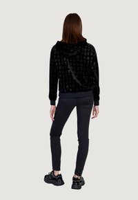 Sudadera con capucha de terciopelo negro con patrón de texto en relieve, combinada con pantalones ajustados negros y zapatillas chunky negras. Vista trasera.