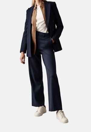 Person iført navyblå jakkesæt med vidde bukser, en tanfarvet skjorte med knapper, hvid top og cremefarvede sneakers, stående med den ene hånd i lommen.