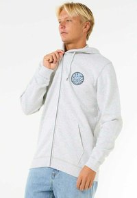 Haut de survêtement zippé gris clair avec capuche à cordons, poches avant et logo graphique circulaire sur la poitrine. Fabriqué à partir d'un tissu doux et texturé.