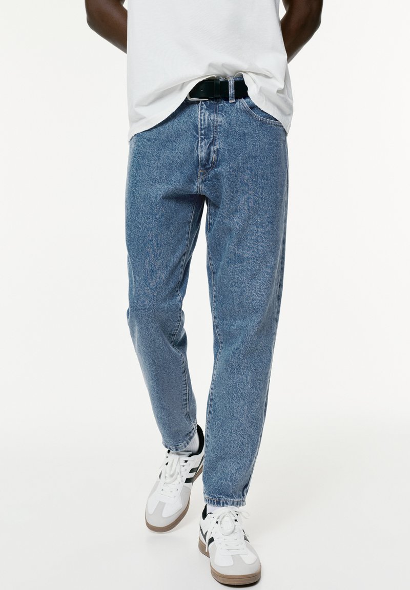 Jeans in denim azzurro chiaro con vestibilità comoda, caratterizzati da un classico design a cinque tasche, indossati con una cintura nera e sneakers bianche.