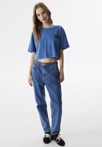 Blaues, kurzes T-Shirt mit einem strukturierten Muster auf der Vorderseite, kombiniert mit lockeren blauen Jeans und schwarzen Turnschuhen mit weißen Schnürsenkeln.
