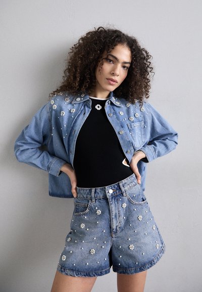 Giacca di denim con decorazioni in perle, indossata sopra una maglia nera. Abbinata a shorts in denim a vita alta con simili accenti in perle.