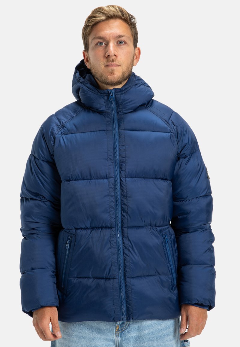 Námornícky modrá páperová bunda so zipsom vpredu, kapucňou a bočnými vreckami. Má prešívaný vzor a hladkú, lesklú textúru.