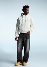 Felpa crema con tasca frontale e design grafico, abbinata a jeans larghi grigio scuro e stivali gialli con silhouette chunky.