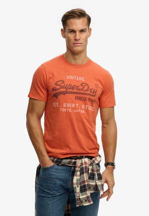 Mann trägt ein oranges Vintage-T-Shirt mit der Aufschrift "SuperDry", blaue Jeans, ein kariertes Hemd um die Taille gebunden und eine Armbanduhr, steht vor einem schlichten Hintergrund.