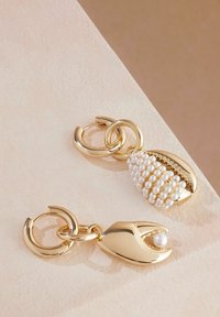 Zlato-tonirana uhani z zakladkom v obliki školjke in s perlovimi detajli ter z zakladkom v obliki kremplja, okrašeni s perlom, na svetli podlagi.