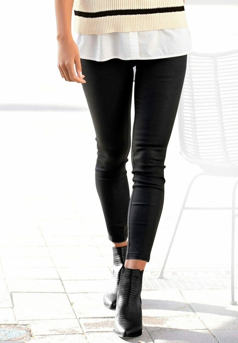 Jambes d'une personne portant un pantalon noir ajusté, des bottines noires texturées, une chemise blanche et un pull beige avec une bande noire.