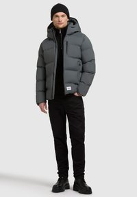 Grijze pufferjack met een capuchon, voorzien van een ritssluiting aan de voorkant en gestikte secties. Gecombineerd met een zwart rits-trui en zwarte broek. Zwarte laarzen.