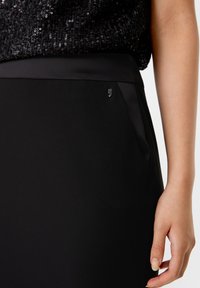 Schwarze Rock mit einer eleganten, glatten Textur; verfügt über ein dezentes Logo-Detail und ein figurbetontes Design. Der Bund ist schlicht und strukturiert.