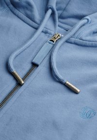 Lichtblauwe hoodie met rits, met metalen ritssluiting en trekkoorden met gegraveerde tekst "SUPERDRY" en subtiel geborduurd logo op de stof.