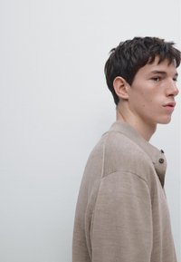 Indumento di un beige chiaro, dalla texture particolare, con colletto a bottoni, caratterizzato da una vestibilità comoda e dettagli a coste sottili, indossato da un giovane uomo.