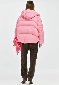 Roze pufferjack met een capuchon, voorzien van gestikte delen. Gecombineerd met donkerbruine broek en een roze franjesaccessoire. Gladde textuur.